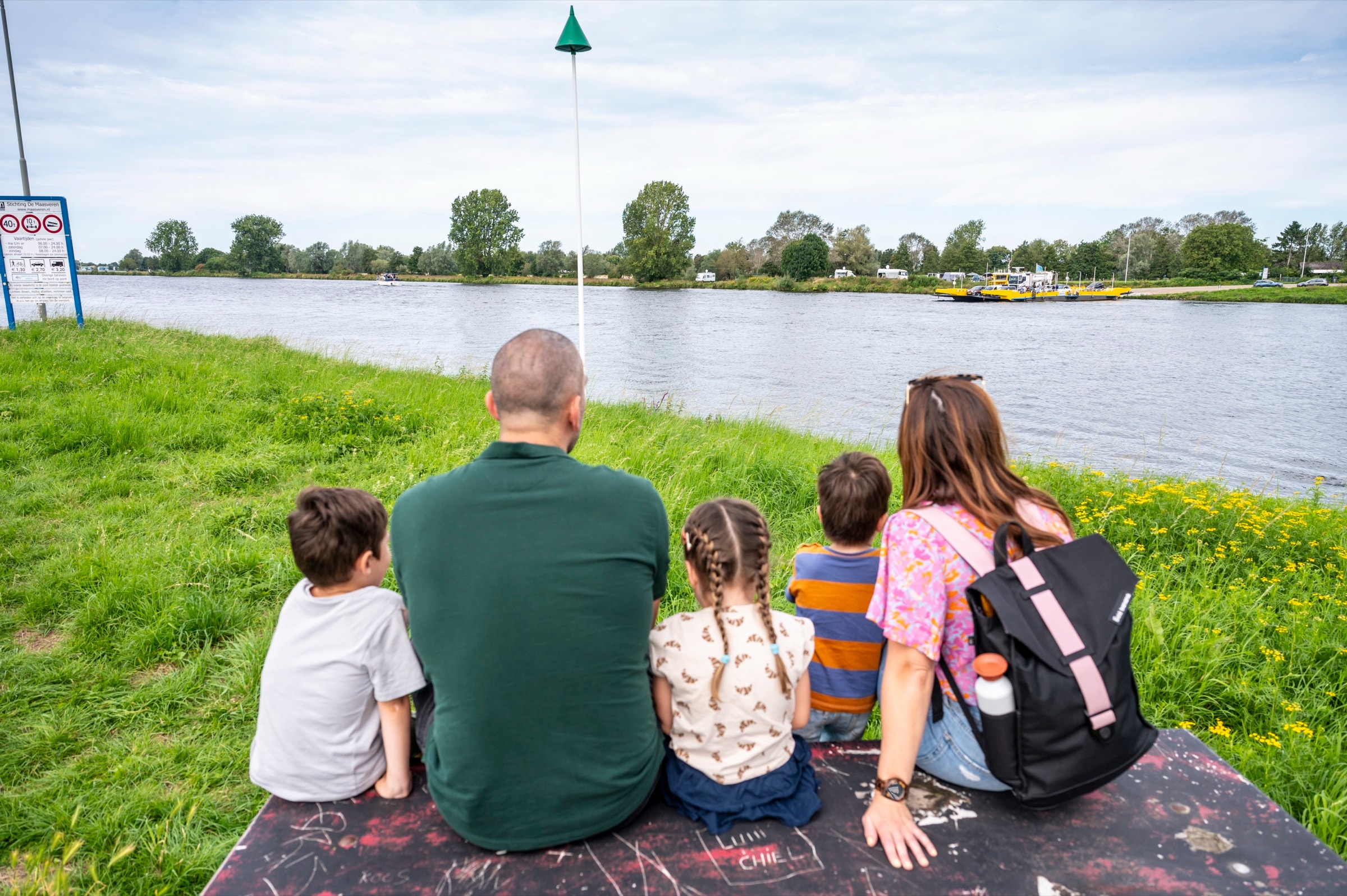 Een gezin met drie kinderen zit op een bank in het gras aan de Maas bij Megen en kijkt over de rivier naar het gele pontje aan de overkant.